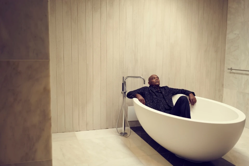 African American male relaxes in a modern bathtub in a bright bathroom with striped walls and marble flooring, wearing a patterned suit.