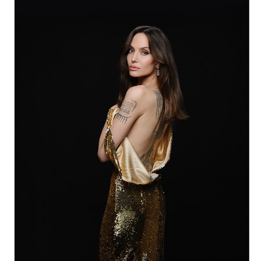 Woman in shimmering gold gown posing against dark backdrop, showcasing elegant jewelry and intricate back tattoos.