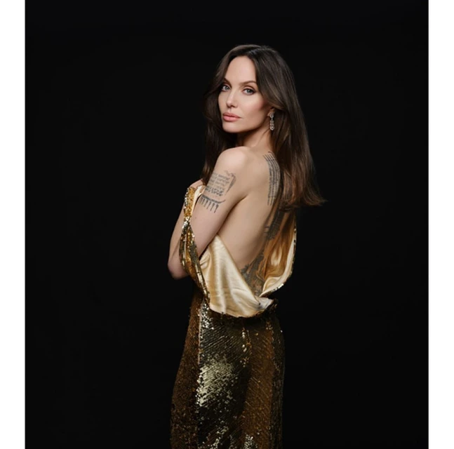 Woman in shimmering gold gown posing against dark backdrop, showcasing elegant jewelry and intricate back tattoos.