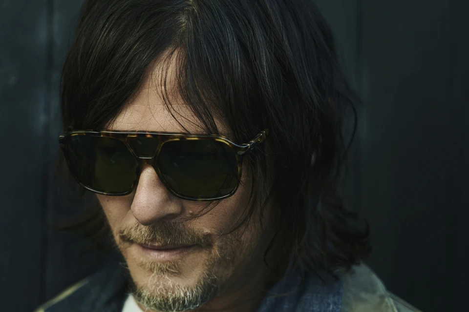 Close-up portrait of an adult male with dark hair and a mustache, wearing sunglasses against a dark blurry background indoors.