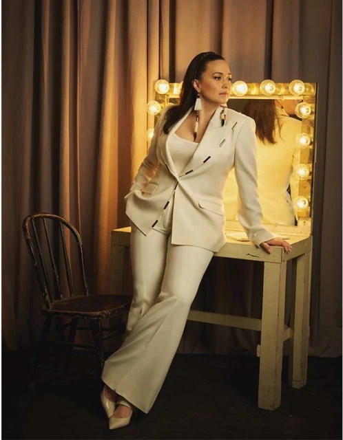 Elegant woman in a cream suit posing indoors next to a vanity mirror and vintage chair, bathed in soft, warm lighting.