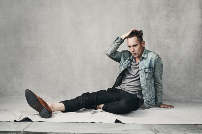 Man in denim jacket and grey t-shirt seated on a grey textured backdrop with one hand in his hair