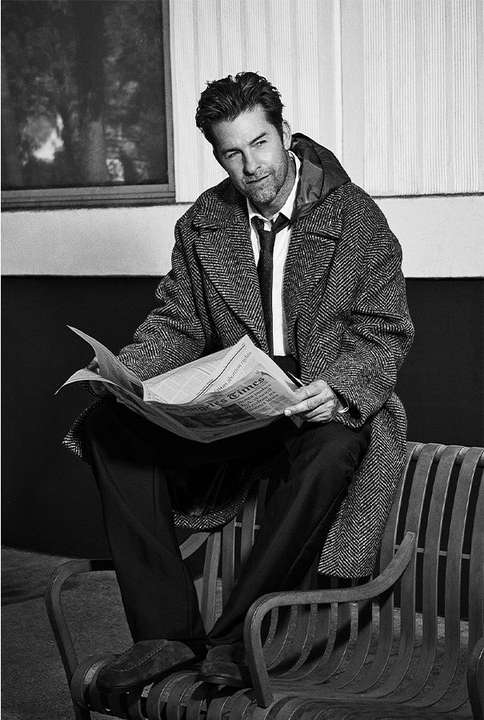 Black and white portrait of a man sitting on a park bench, reading a newspaper, wearing an overcoat and tie outdoors.