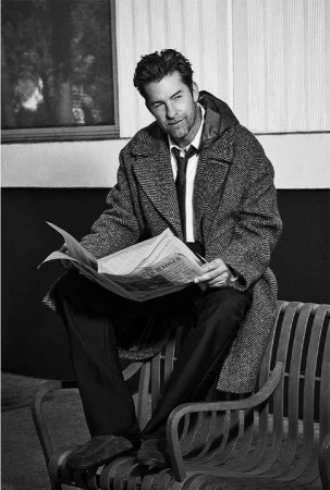 Black and white portrait of a man sitting on a park bench, reading a newspaper, wearing an overcoat and tie outdoors.