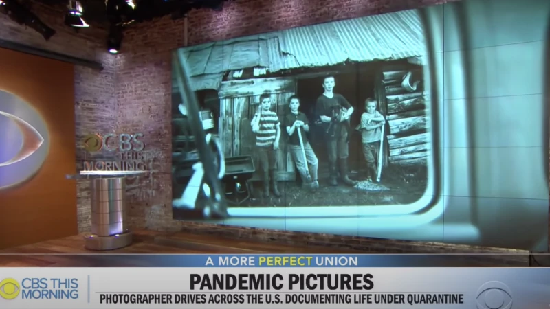 Black and white photograph of five children standing in front of a log cabin is displayed on a monitor within a television studio.