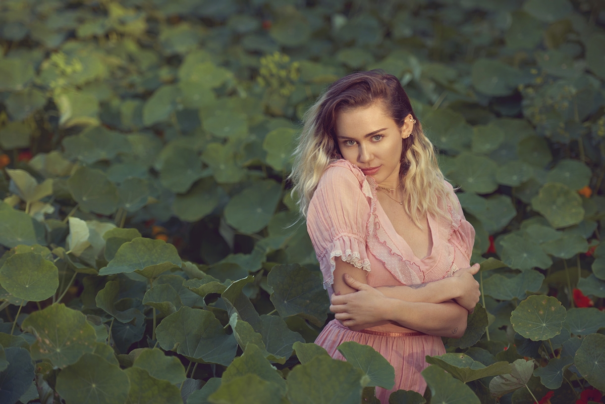 Blonde woman in pink dress surrounded by green leaves in a natural outdoor setting, gazing directly at the camera with soft, diffused lighting.