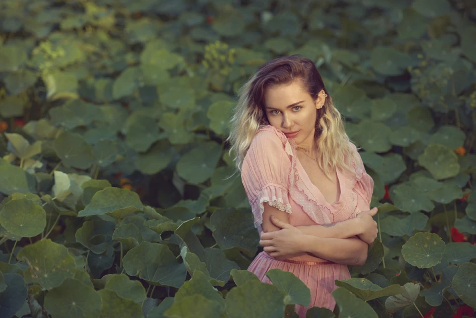 Blonde woman in pink dress surrounded by green leaves in a natural outdoor setting, gazing directly at the camera with soft, diffused lighting.