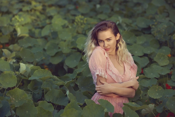 Blonde woman in pink dress surrounded by green leaves in a natural outdoor setting, gazing directly at the camera with soft, diffused lighting.