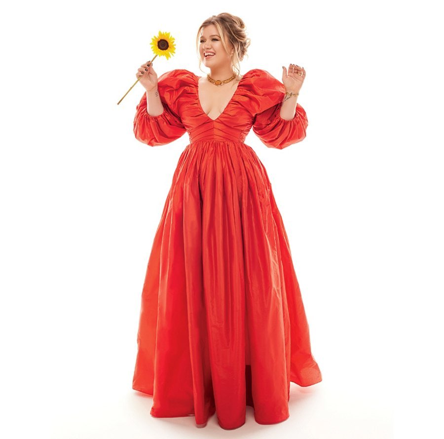 Kelly Clarkson holding a sunflower wearing a long red puffy-sleeved dress against a white background