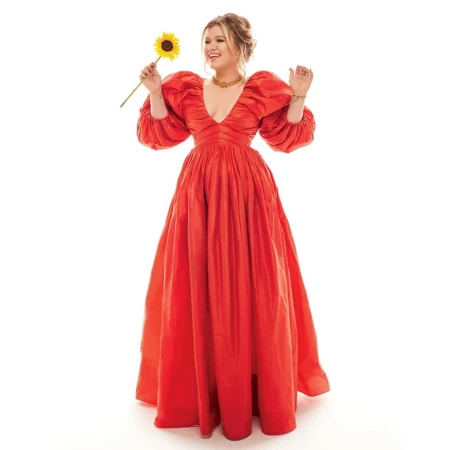 Kelly Clarkson holding a sunflower wearing a long red puffy-sleeved dress against a white background