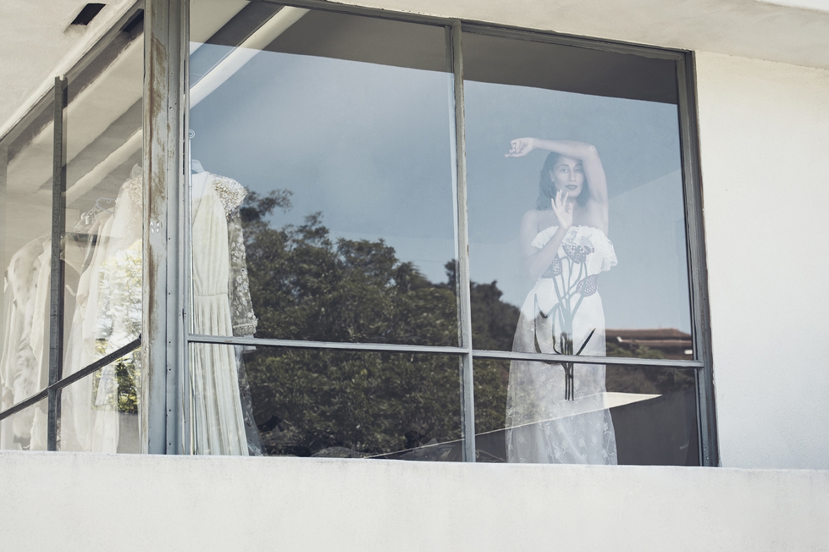 Woman in white lace dress reflected in window with wedding dresses, outdoor setting, daylight, clear sky, eye-level perspective.