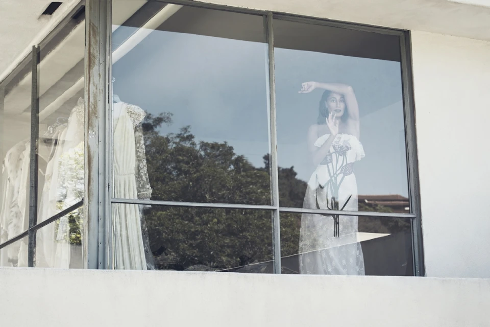Woman in white lace dress reflected in window with wedding dresses, outdoor setting, daylight, clear sky, eye-level perspective.