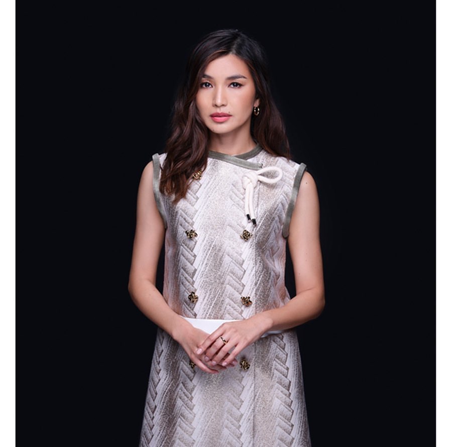 Portrait of a woman with long brown hair, wearing a patterned dress with decorative buttons, posing against a dark grey background.