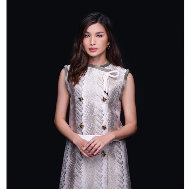 Portrait of a woman with long brown hair, wearing a patterned dress with decorative buttons, posing against a dark grey background.
