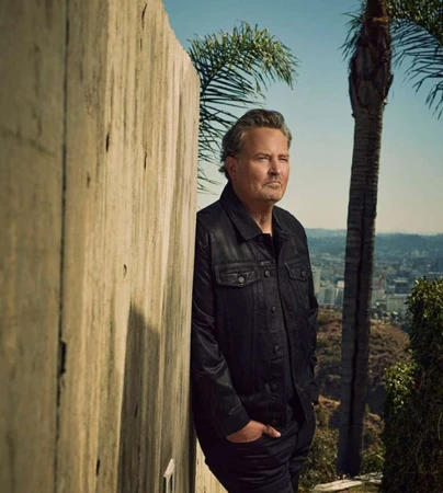 Matthew Perry leaning against a concrete wall wearing a dark jacket with palm trees and a city view behind him.