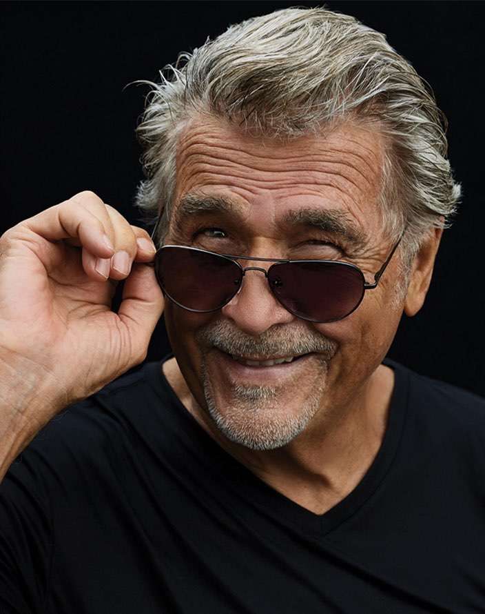 Close-up portrait of an older man with salt and pepper hair wearing sunglasses, smiling against a black background.