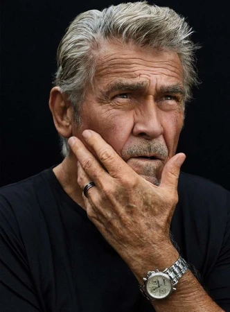 Portrait of an older man with silver hair and a mustache wearing a black t-shirt against a dark background