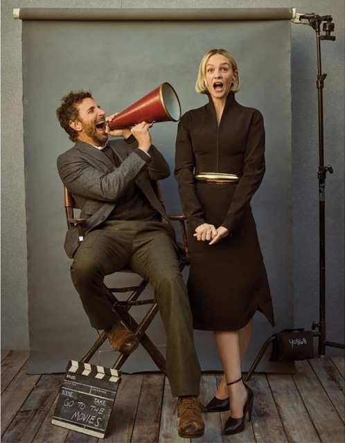 Man and woman pose with a megaphone and film slate in a photo studio, showcasing vintage fashion and cinematic style with a gray backdrop.