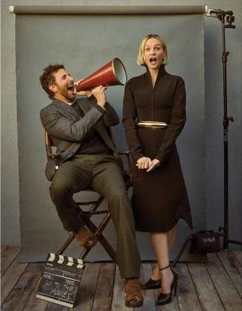 Man and woman pose with a megaphone and film slate in a photo studio, showcasing vintage fashion and cinematic style with a gray backdrop.