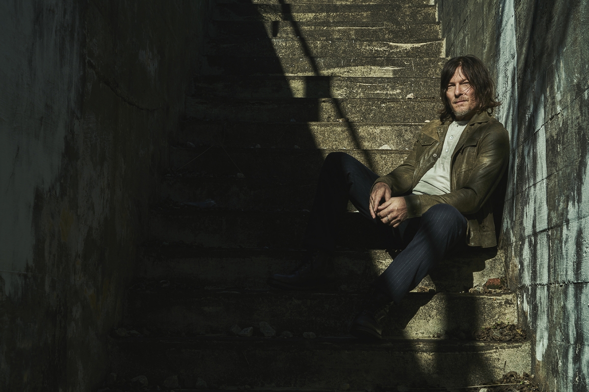 Adult man with long hair sitting on a concrete staircase wearing a jacket, jeans, and henley shirt in shadows and light.