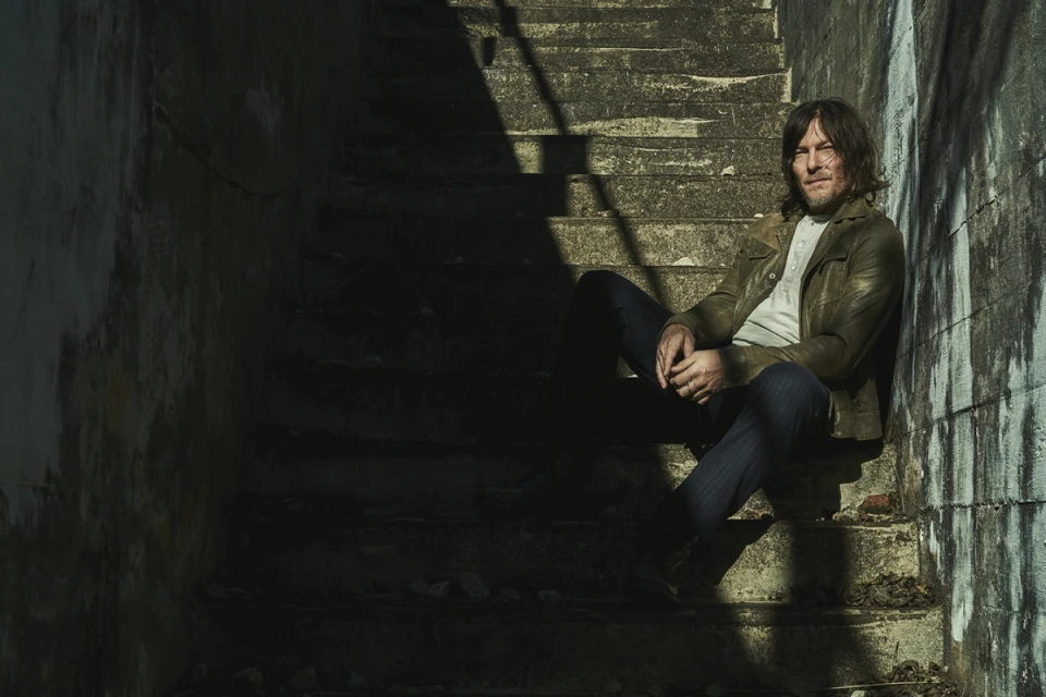 Adult man with long hair sitting on a concrete staircase wearing a jacket, jeans, and henley shirt in shadows and light.