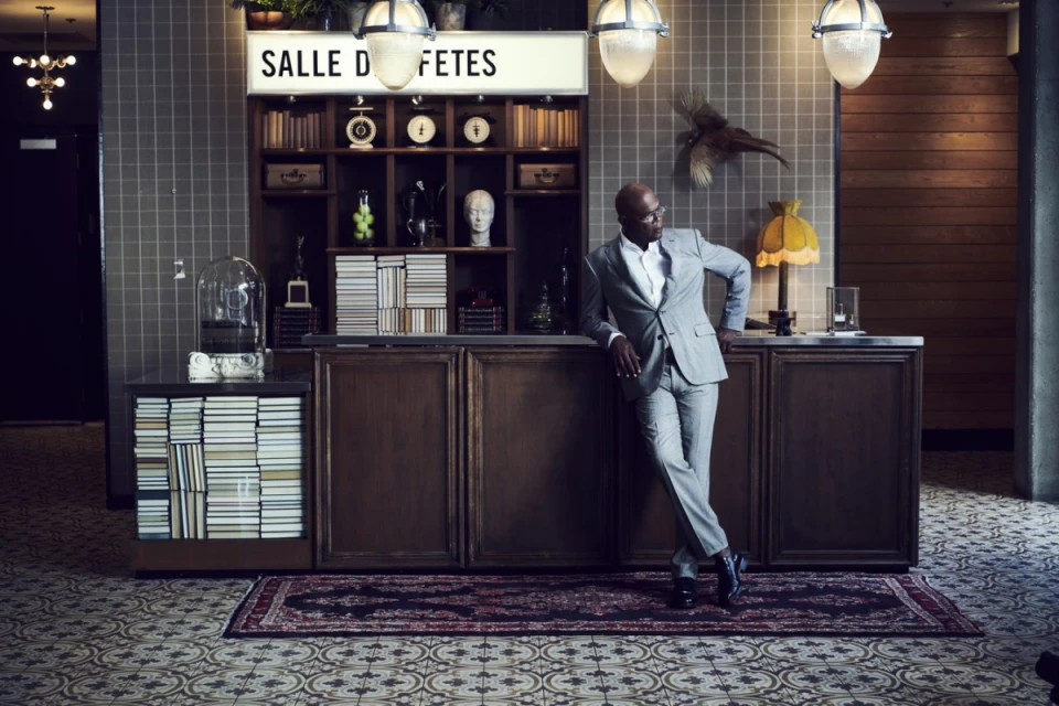 Well-dressed Black man in light grey suit posing in ornate interior with tile floors, books, desk, and a 'Salle des Fetes' sign above.