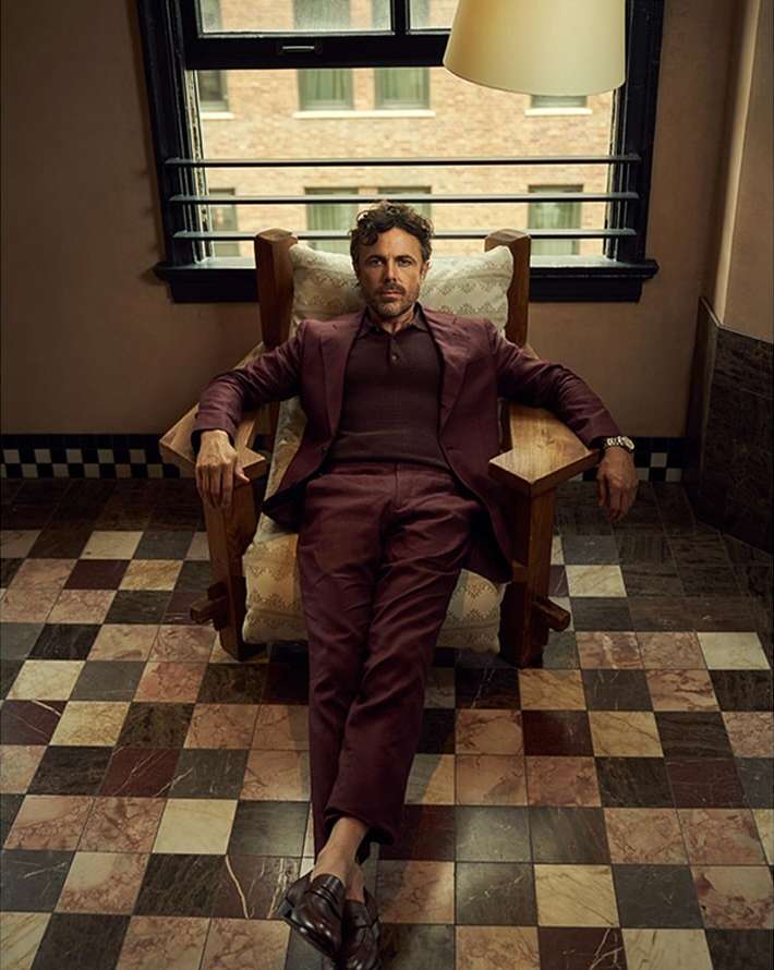 A man in a maroon suit sits in a wood chair in an indoor setting with a patterned tile floor and light coming from a window.