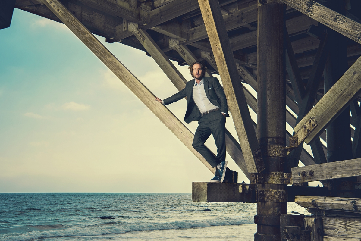 Man wearing a dark suit and sneakers standing under a wooden pier structure above the ocean, photographed by Brian Bowen Smith.