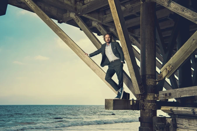 Man wearing a dark suit and sneakers standing under a wooden pier structure above the ocean, photographed by Brian Bowen Smith.