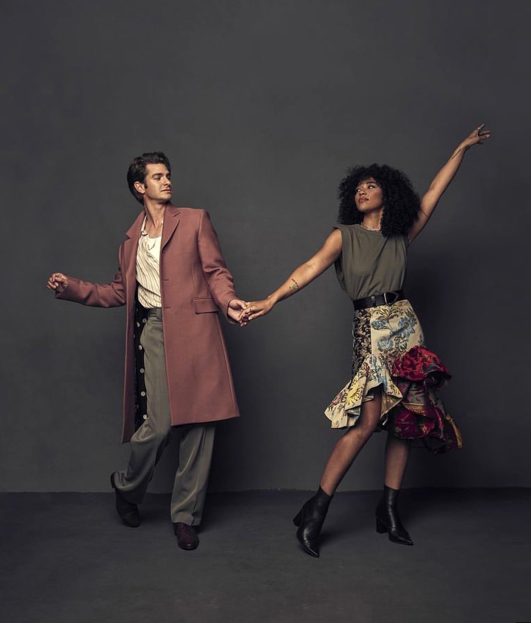 Andrew Garfield and Alexandra Shipp pose together holding hands in a studio. Garfield wears a long coat, Shipp wears a skirt and boots.