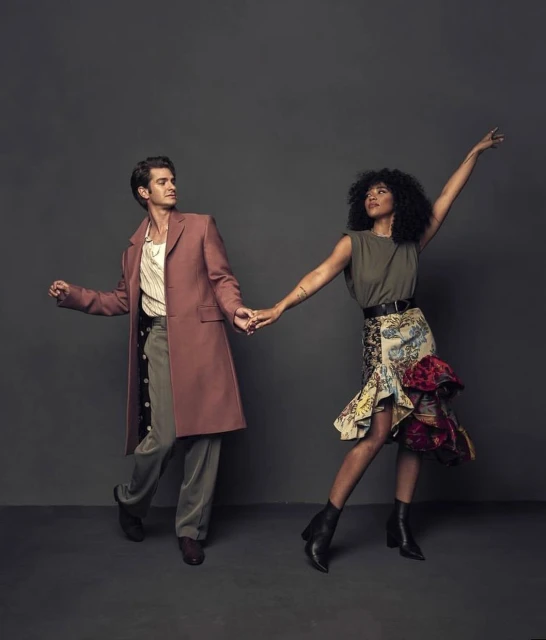 Andrew Garfield and Alexandra Shipp pose together holding hands in a studio. Garfield wears a long coat, Shipp wears a skirt and boots.