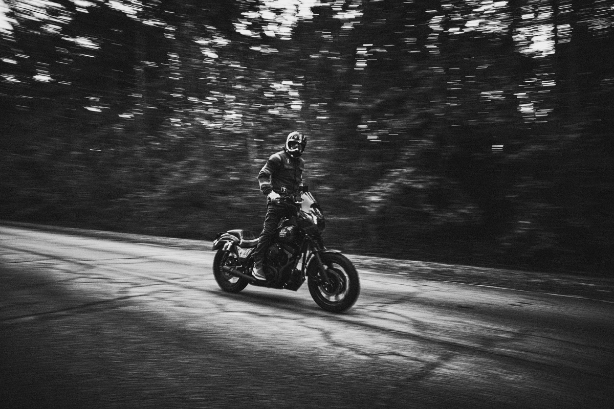 Monochrome action shot of a motorcyclist riding a black motorbike on an asphalt road through a forest; motion blur indicates speed.