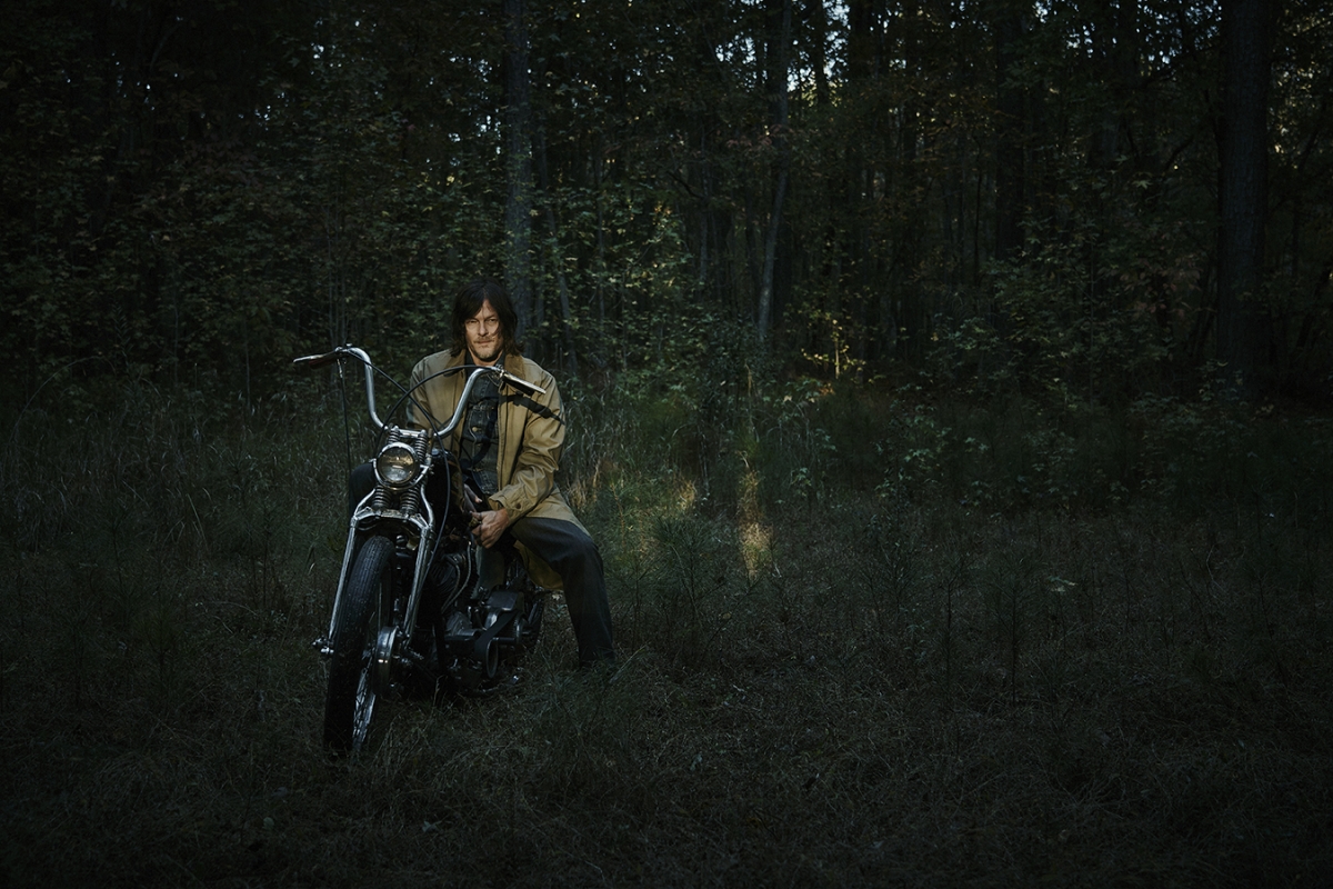 Adult male poses on a black motorcycle in a forest setting, wearing a tan trench coat and dark trousers on an overcast day.