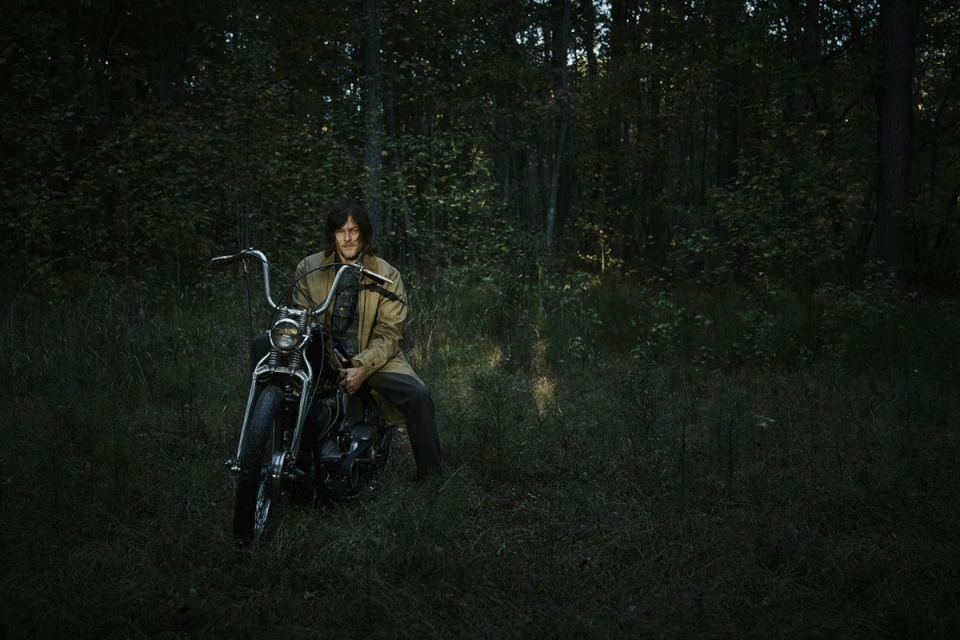 Adult male poses on a black motorcycle in a forest setting, wearing a tan trench coat and dark trousers on an overcast day.