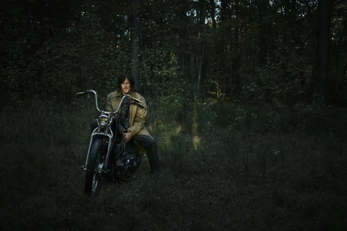 Adult male poses on a black motorcycle in a forest setting, wearing a tan trench coat and dark trousers on an overcast day.