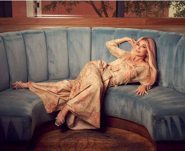 Shania Twain, reclining on a blue velvet sofa, wears a patterned wide-leg suit with silver heeled sandals in a softly lit interior.