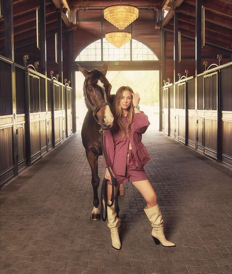 A woman poses with a horse in an indoor stable, wearing a pink shorts suit and cream-colored boots, chandeliers are overhead.