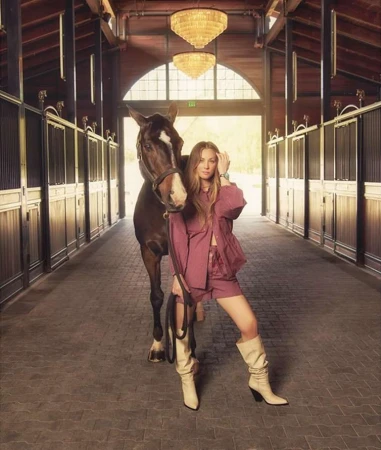 A woman poses with a horse in an indoor stable, wearing a pink shorts suit and cream-colored boots, chandeliers are overhead.