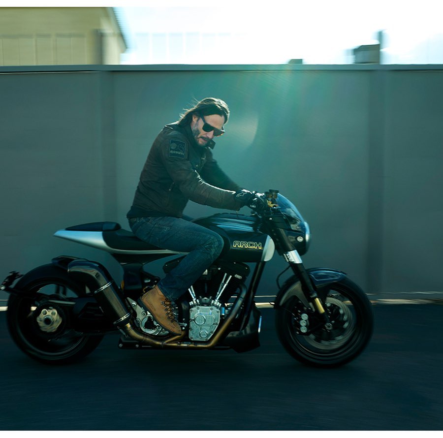 Keanu Reeves rides a ARCH motorcycle, wearing a jacket, jeans, and boots. The bike is black and silver, set against an urban backdrop.
