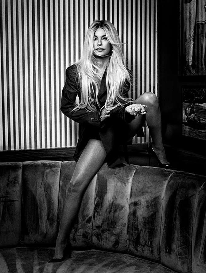 Black and white portrait of a blonde woman in an oversized blazer sitting on a tufted curved booth against striped wallpaper