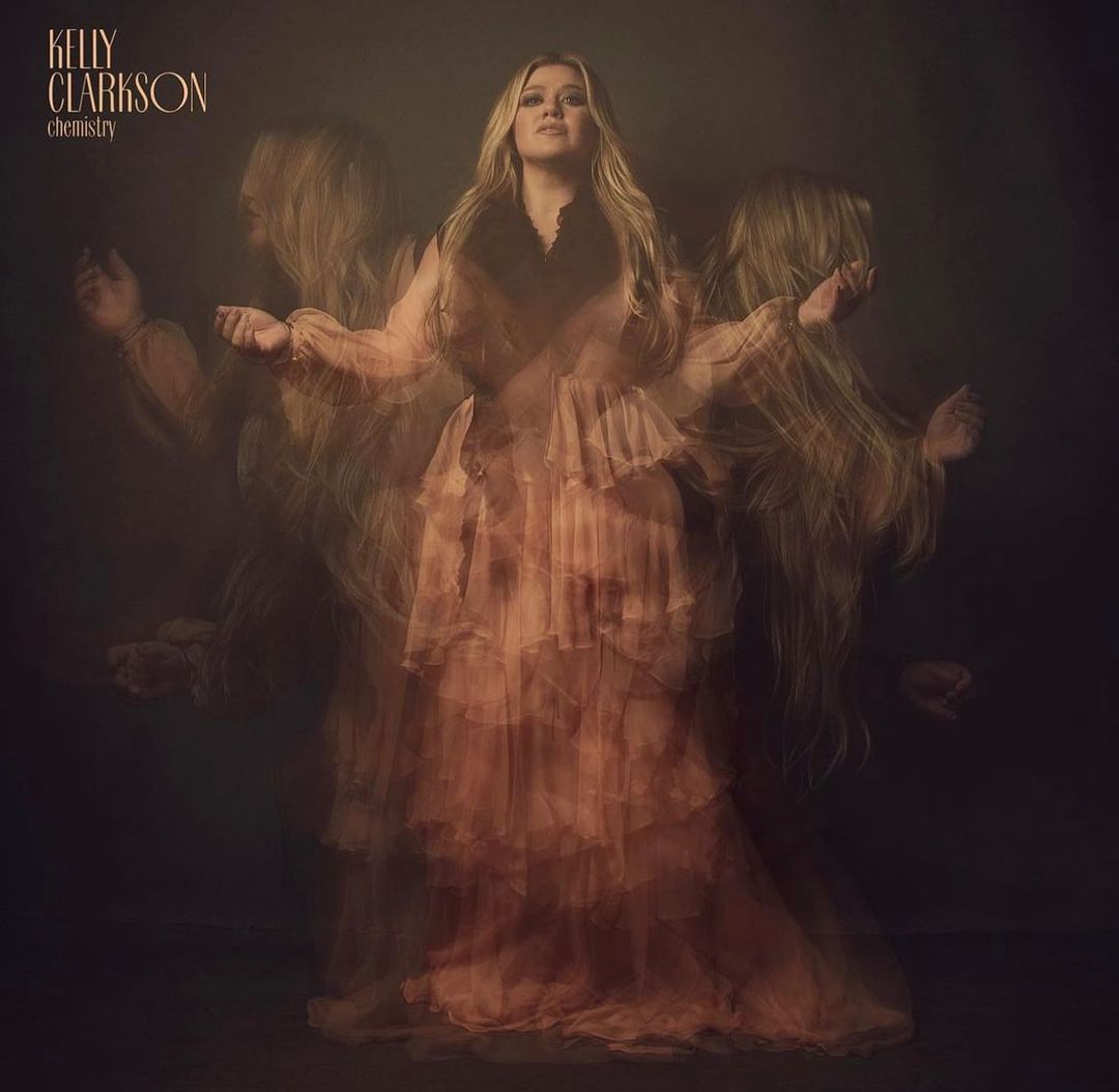 Kelly Clarkson poses in a peach-colored tiered tulle gown, captured in a blurred, long exposure style with a dark moody background.