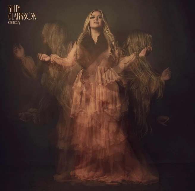 Kelly Clarkson poses in a peach-colored tiered tulle gown, captured in a blurred, long exposure style with a dark moody background.