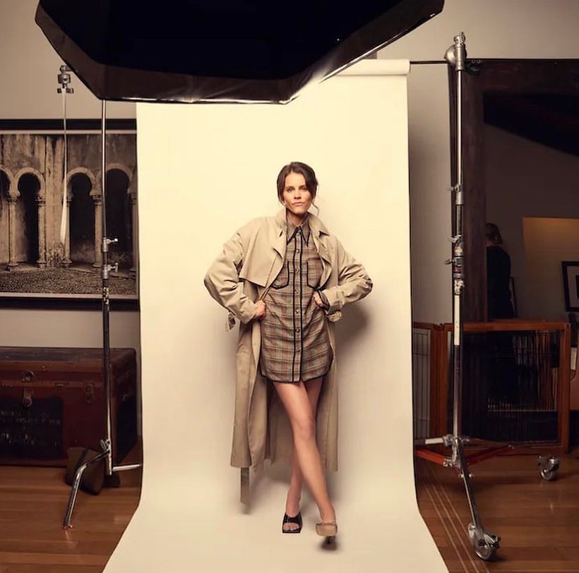 Adult woman in studio, wearing beige trench coat and plaid shirt dress, poses in front of white backdrop with studio lighting and equipment.