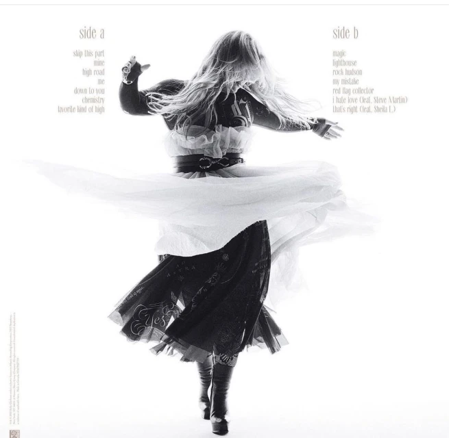 A woman wearing a black-and-white tulle dress with long blonde hair dances with her arms outspread on a white background in a studio shot.