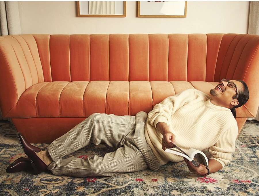 Man lounges on rug near an orange sofa, engrossed in a book and smiling, wearing glasses and a cream sweater in an indoor setting.