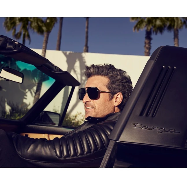 Adult man with sunglasses driving a black convertible Targa under palm trees with a blue sky, wearing a leather jacket.