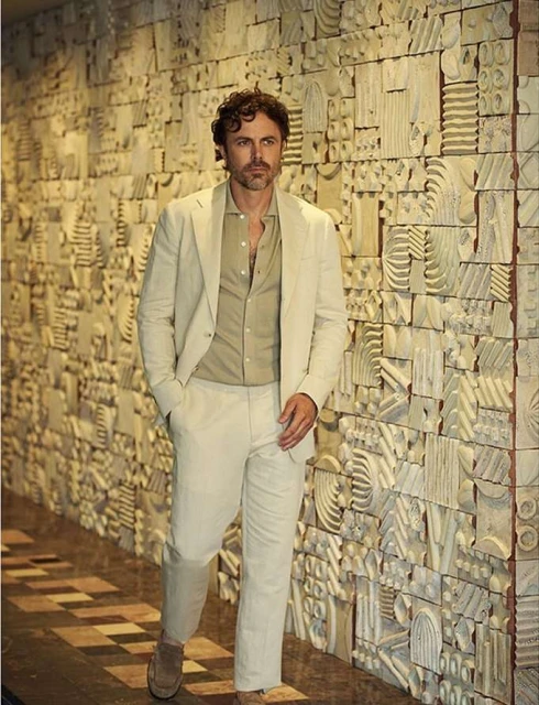 Adult male in cream suit poses against an intricately designed wall, adding depth and sophistication to the fashion shot.
