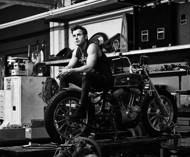 Black and white portrait of a man sitting on a motorcycle in a cluttered workshop, looking off to the side with a serious expression.