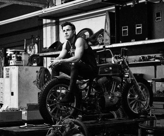 Black and white portrait of a man sitting on a motorcycle in a cluttered workshop, looking off to the side with a serious expression.