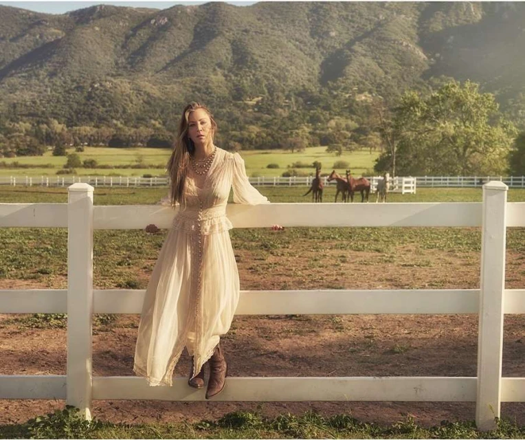 Woman in long dress leans on white fence with horses and mountain backdrop, creating a serene, pastoral aesthetic.